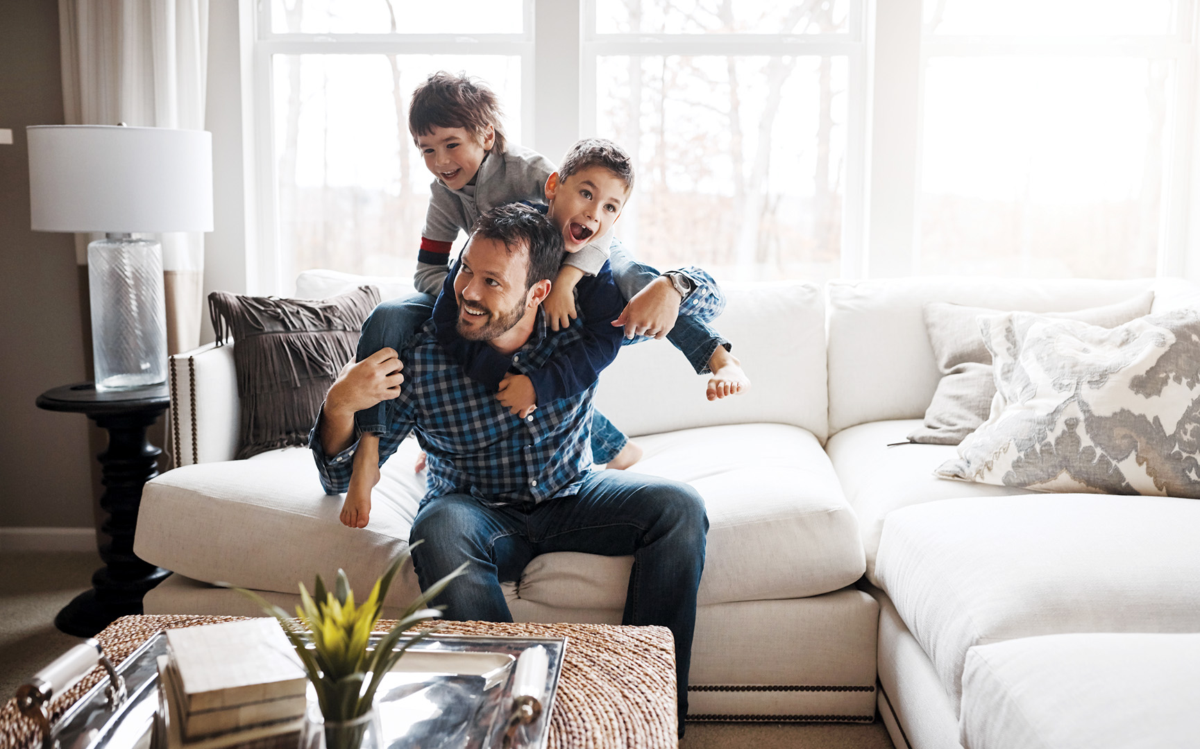 Rough and tumble play when raising boys helps to diffuse excess emotional energy. Two young boys are playfully on their father's back. Father is sitting on a couch in the living room.