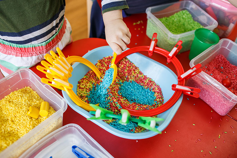 Indoor activities for kids. Toddlers playing with sensory bin with colourful rice on red table.