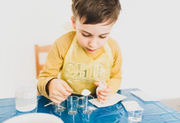 Toddler at table pouring spoonful of white powder into a small glass. things to do with kids - science experiments