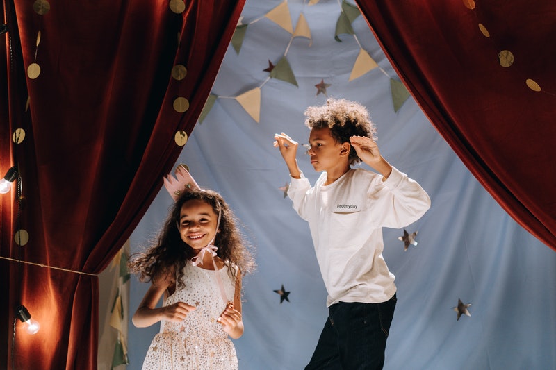 A young boy and girl in action in front of a homemade stage backdrop. The young girl is wearing a homemade crown and smiling. The young boy has arms raised and is in mid-jump. Things to do with kids - storytelling