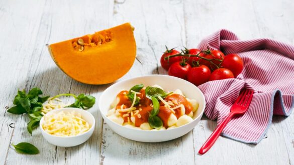 Bowl of gnocchi on table flanked by pumpkin, tomatoes and cheese