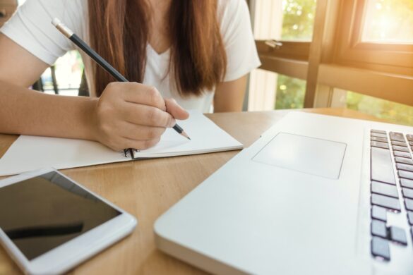 torso image of girl sitting, about to write in blank book with a pencil. there is a laptop and a tablet in the foreground