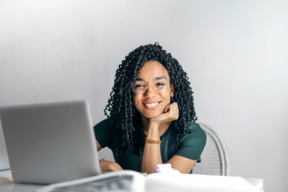 Black woman with glasses sitting in front of a laptop, smiling at the camera