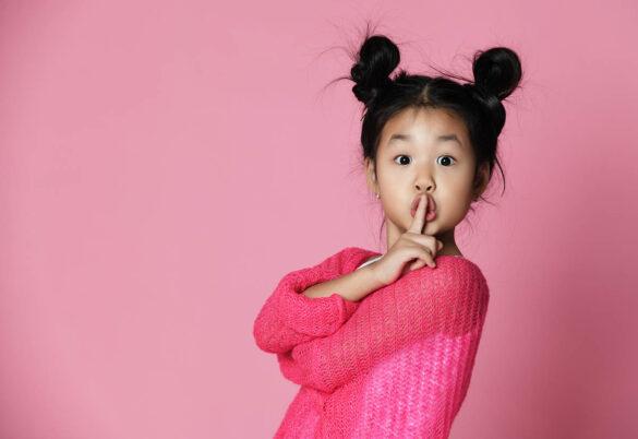 Asian kid girl in pink sweater shows shh sign on pink background. Close up portrait