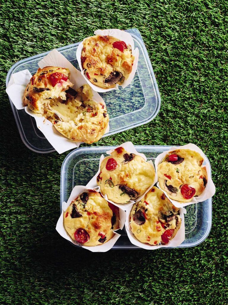 Six vegetable muffins spread over two clear plastic containers. In one container, there are two muffins with one muffin split open to show how it looks inside.