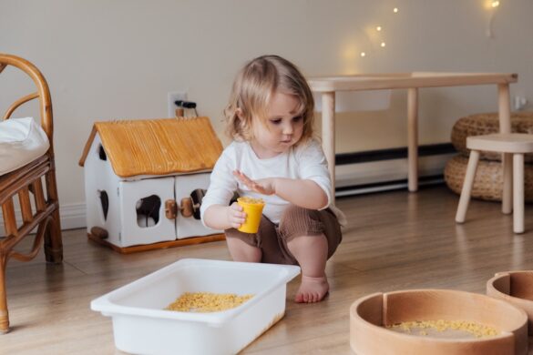 Toddler squatting in a living room holding a yellow plastic cup full of dried macaroni
