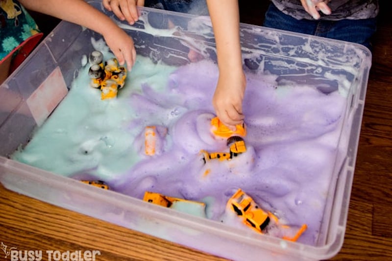Toddler hands reaching into a low plastic tub filled with green and purple foam, playing with toy construction cars.
