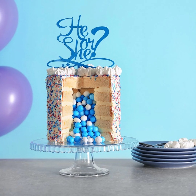Five layer sponge cake with a large slice cut out, revealing the middle of the cake. Little blue and white balls have tumbled out from the inside. Cake topper says he or she? 