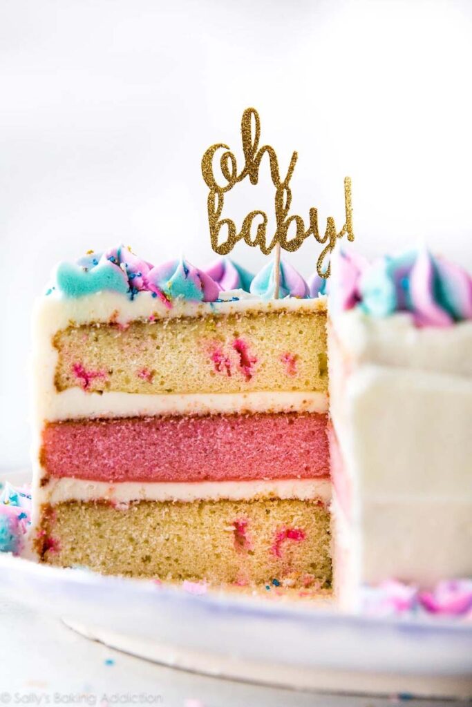 Side view of a slice of three layer sponge cake. The top and bottom layer is a pale yellow while the middle layer is pink. Cake topper says oh baby!