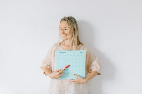 Cycle syncing - Smiling woman holding a monthly calendar in front of her standing against a white background. She is pointing to a date on the calendar with a red marker pen.