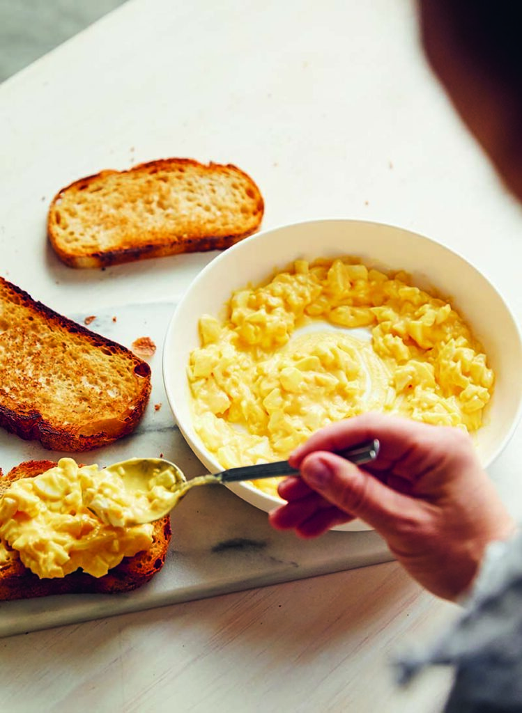 Bowl of creamy egg salad in a bowl with three slices of toast on the table. A hand has scooped out a spoonful of egg salad to spread over a slice of toast.