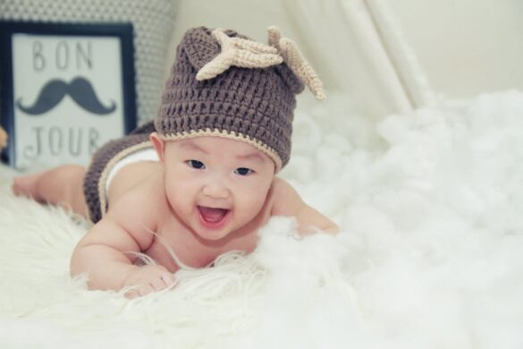 Baby grinning widely and lying on stomach on a white fluffy blanket wearing a crocheted beanie and nappy cover