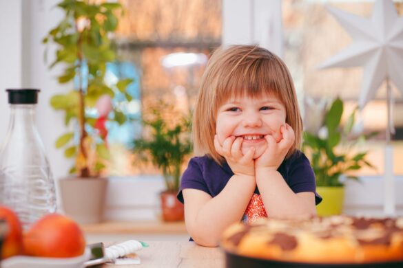 Smiling child resting head on hands, with elbows on kitchen bench.