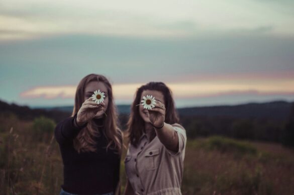 Two smiling women at dusk standing in a field holding up a daisy each in front of their faces