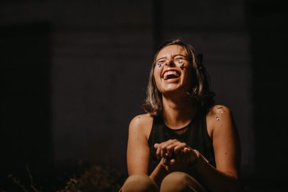 Woman with a wide smile sitting in front of a dark background with glitter confetti pieces stuck on her body and face