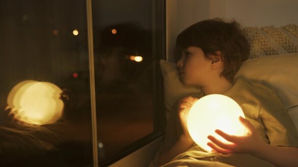 Boy sitting in bed by the window. It's at night, he's holding a ball of light and looking out the window.